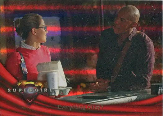Two people conversing at a desk with colorful lighting and a Supergirl Rainbow Foil Board