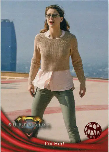 Woman in glasses with a beige sweater and green pants beside Supergirl Red Omegahedron Deco